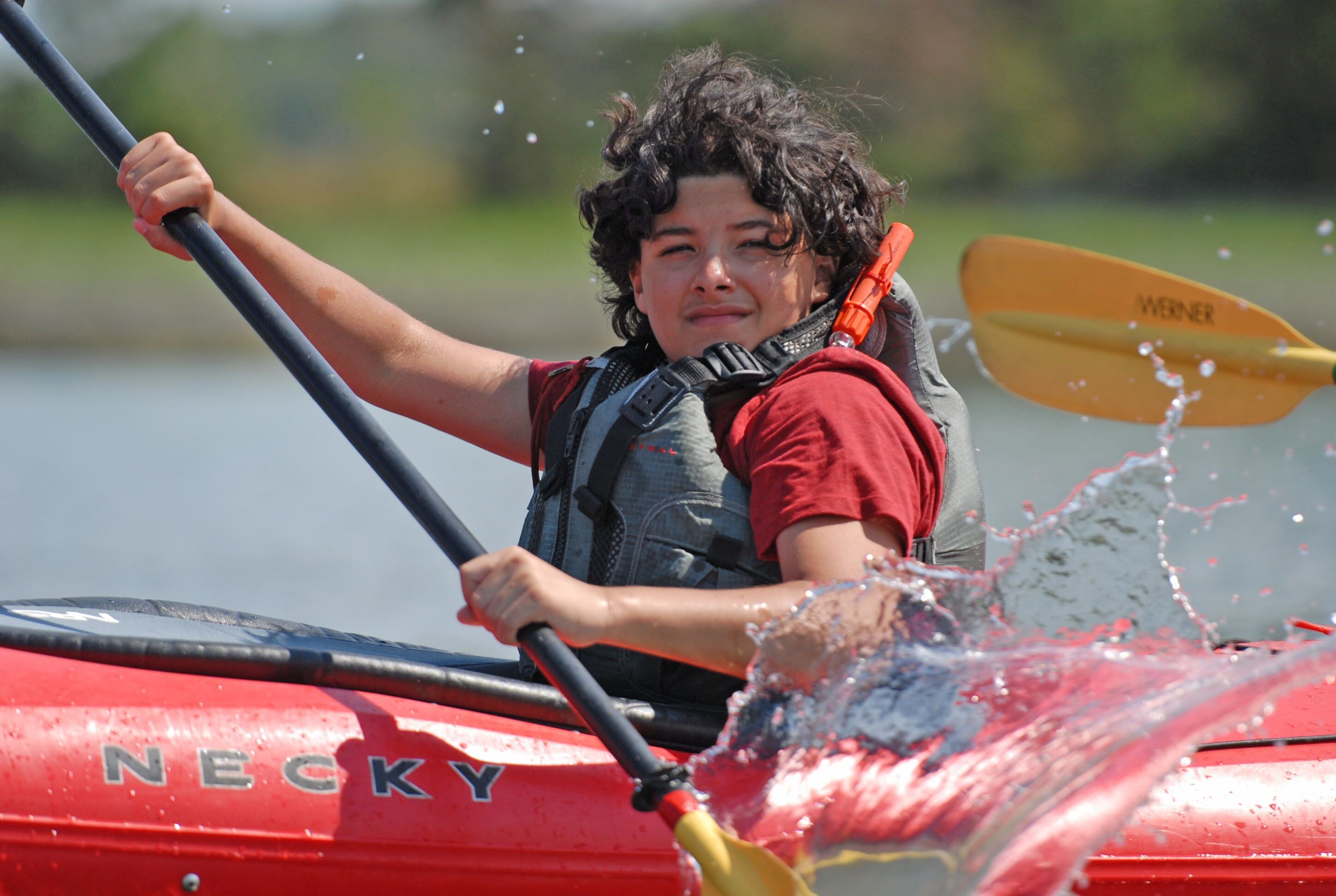 The image shows a young person kayaking on a sunny day. They are wearing a life vest and appear to be enjoying themselves. The kayak is red, and the water is splashing around the paddle. The background is blurred, suggesting a focus on the kayaker and the action.